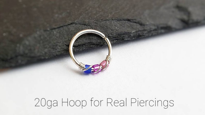 Puede incluir: Un pendiente de aro plateado con cuentas azules, rosas y moradas. El texto "20ga Hoop for Real Piercings" est&aacute; en la parte inferior de la imagen. El pendiente est&aacute; sobre una superficie gris oscuro.
