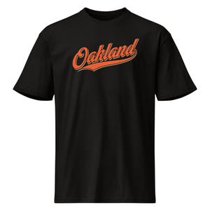 Könnte beinhalten: Schwarzes T-Shirt mit dem Wort "Oakland" in orangefarbener Schrift. Der Text hat einen Schatteneffekt. Das T-Shirt ist im Rundhals-Stil, ein gängiges Design für Freizeitkleidung.