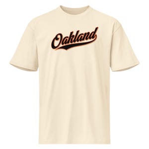 Könnte beinhalten: Cremefarbenes T-Shirt mit dem Wort "Oakland" in Schreibschrift. Der Text ist schwarz umrandet und hat einen rot-orangen Schatten. Das Shirt hat kurze Ärmel und einen Rundhalsausschnitt.