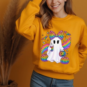 May include: A mustard yellow long-sleeve sweatshirt featuring a central graphic of a white ghost with a rainbow leopard print bow, rainbow wings, and a drink. The ghost is surrounded by two rainbow leopard print pumpkins, bats, and stars.