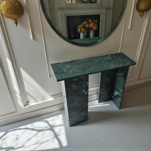Puede incluir: Una consola de mármol verde con una tapa rectangular y dos patas de soporte. La mesa está hecha de mármol verde oscuro con vetas blancas. La mesa está colocada contra una pared blanca.
