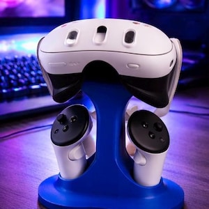 May include: A white and black VR headset and controllers on a blue stand. The stand is on a wooden surface. The VR headset has multiple small openings on the top.