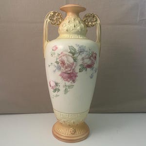 Op de afbeelding: Crèmekleurige keramische vaas met roze rozenbloemmotief. De vaas heeft twee handvatten met gouden accenten en een decoratieve voet. De bovenkant en de voet zijn licht perzikkleurig. Een klassiek decoratief stuk.