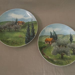 Puede incluir: Dos platos decorativos redondos con una escena de paisaje pintada. Cada plato presenta una casa con techo naranja, árboles y colinas verdes onduladas bajo un cielo azul con nubes. Los platos tienen un borde blanco.