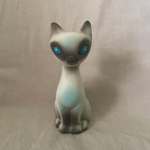 Op de afbeelding: Een vintage Siamese kattenbeeldje in lichtblauwe en bruine tinten. De kat heeft helderblauwe ogen en zit. Het beeldje is waarschijnlijk gemaakt van keramiek of een vergelijkbaar materiaal.