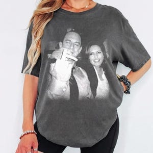 Pode incluir: T-shirt cinzenta escura com uma imagem a preto e branco de duas pessoas. A imagem está centrada na frente da t-shirt. A t-shirt tem mangas curtas e gola redonda. A t-shirt é usada por uma pessoa.
