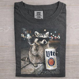 May include: Dark gray t-shirt featuring a raccoon wearing sunglasses and smoking a cigarette, holding a can of Lite beer. The shirt has a Comfort Colors tag.