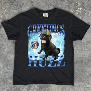 May include: A black short-sleeve t-shirt featuring a graphic of a black pug wearing a colorful propeller beanie. The dog is next to a multi-colored lollipop against a blue lightning background. The text GREETINGS and HUZZ is printed in blue.