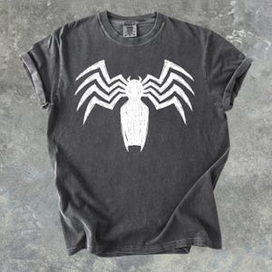 Könnte beinhalten: Dunkelgraues T-Shirt mit einem weißen, von Venom inspirierten Motiv. Das Design ist ein stilisierter spinnenartiger Umriss mit scharfen, kantigen Linien. Das T-Shirt hat hochgekrempelte Ärmel und einen Rundhalsausschnitt.