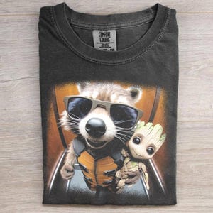 May include: Dark grey t-shirt with a graphic of Rocket Raccoon in sunglasses and Groot. The shirt has a crew neck and the Comfort Colors brand label is visible. The design is printed on a folded shirt.
