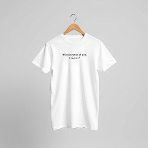 Puede incluir: Camiseta blanca con el texto "Min partner är bra. I teorin." en negro. La camiseta está colgada de una percha de madera sobre un fondo gris claro. La camiseta es de cuello redondo.