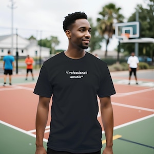 Puede incluir: Camiseta negra con el texto "Professionell amatör" en blanco. La camiseta la lleva una persona en una cancha de baloncesto. Hay otras personas visibles en el fondo.