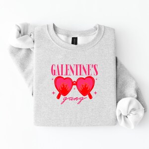 May include: A light gray sweatshirt with the text "GALENTINE'S" above a pair of pink heart-shaped sunglasses with flames and the word "gang" below. The sleeves are rolled up.