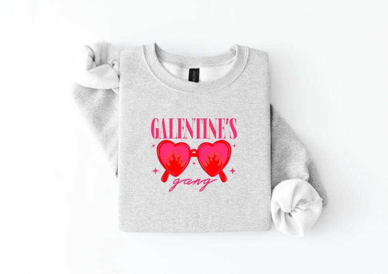 May include: A light gray sweatshirt with the text "GALENTINE'S" above a pair of pink heart-shaped sunglasses with flames and the word "gang" below. The sleeves are rolled up.