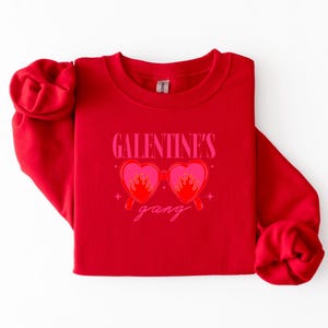 May include: A red sweatshirt with the text "GALENTINE'S gang" in pink. The design features heart-shaped sunglasses with flames. The sweatshirt is folded on a white surface.