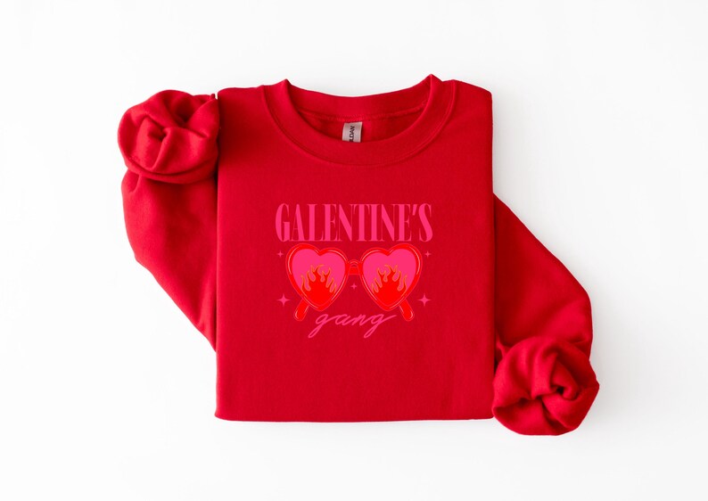 May include: A red sweatshirt with the text "GALENTINE'S gang" in pink. The design features heart-shaped sunglasses with flames. The sweatshirt is folded on a white surface.