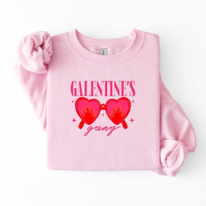 May include: A light pink sweatshirt with the text "GALENTINE'S gang" in pink script. The design features heart-shaped sunglasses with flames. The sweatshirt is folded and displayed on a white surface.