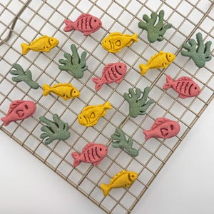 May include: A collection of small, textured fish and coral shapes in shades of pink, yellow, and green. The fish are detailed with etched lines for fins and eyes, while the coral pieces have branching forms. These items are arranged on a metal grid.