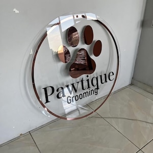 Puede incluir: Un letrero circular transparente con un borde de oro rosa y un diseño de huella de pata de oro rosa. El letrero dice "Pawtique Grooming" en letras negras. El letrero está montado en una pared blanca.