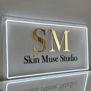 May include: A rectangular sign with a white border and the text "SM Skin Muse Studio." The letters "SM" are gold, and the text is black. The sign is illuminated with a white light.