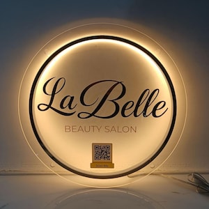 May include: A round, illuminated sign for "La Belle Beauty Salon." The sign features a black border, a warm light, and the salon's name in elegant script. Below, it reads "BEAUTY SALON" and includes a QR code with "Scan Me."