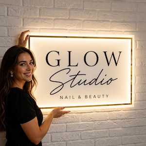 Può includere: Un'insegna rettangolare a LED montata a parete con cornice dorata e sfondo bianco illuminato. L'insegna presenta il testo nero GLOW in carattere serif, Studio in carattere corsivo e NAIL & BEAUTY in carattere sans-serif in basso.