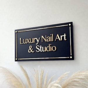 Op de afbeelding: Een marineblauw rechthoekig bord met gouden letters met de tekst "Luxury Nail Art & Studio". Het bord heeft een gouden rand en is bevestigd aan een witte muur. Het is een decoratief bord voor een nagelsalon.