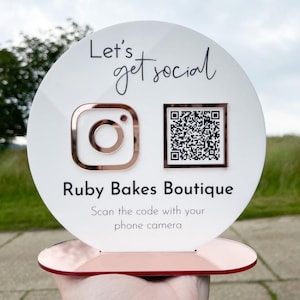May include: A white and rose gold sign with the text "Let's get social" and "Ruby Bakes Boutique". The sign features an Instagram logo and a QR code. The base of the sign is rose gold, and the text "Scan the code with your phone camera" is also included.