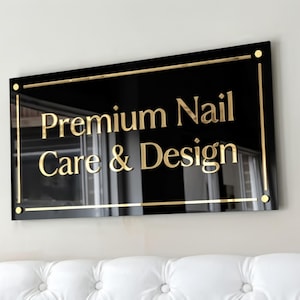 Peut inclure: Une enseigne rectangulaire noire avec des lettres dorées indiquant Premium Nail Care & Design. L'enseigne présente une fine bordure dorée et quatre accents circulaires dorés à chaque coin, fixée sur un mur au-dessus d'une surface capitonnée blanche.