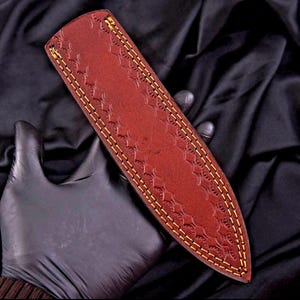 May include: A brown leather knife sheath with a decorative diamond pattern and yellow stitching. The sheath is designed to protect a knife blade. The leather has a rich, reddish-brown color.