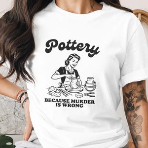 Op de afbeelding: Wit t-shirt met het woord "Pottery" in een retro lettertype boven een zwart-wit illustratie van een vrouw die aan een pottenbakkerswiel werkt. De tekst "BECAUSE MURDER IS WRONG" staat eronder.