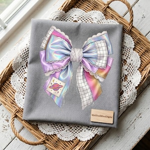 May include: A gray fabric item featuring a large, colorful bow illustration. The bow has iridescent, purple, and pink ribbons with white lace trim. A small patch with a planet design is visible. The item is on a wicker tray.