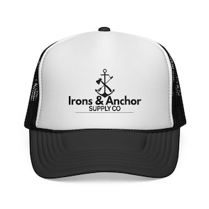 May include: A black and white trucker cap with a curved peak. The white front panel displays a black logo of an anchor, axe, and the text "Irons & Anchor SUPPLY CO". The cap has a black mesh back and a black peak.