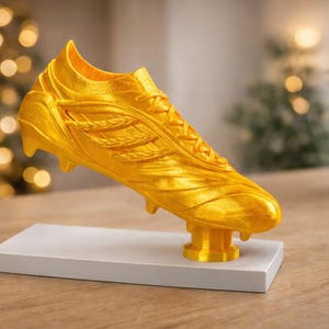 May include: A golden soccer cleat sculpture, mounted on a white rectangular base. The cleat is detailed with laces and textured panels. The background features a blurred bokeh effect and a potted plant.