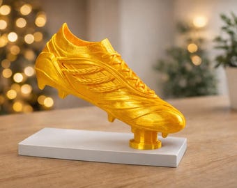 May include: A golden soccer cleat sculpture, mounted on a white rectangular base. The cleat is detailed with laces and textured panels. The background features a blurred bokeh effect and a potted plant.