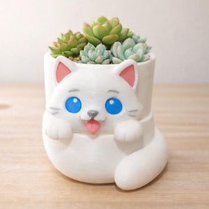Puede incluir: Maceta blanca con forma de gato con diseño juguetón, con ojos azules, orejas rosas y lengua rosa. Contiene varias suculentas verdes, creando una pieza decorativa encantadora para el hogar o la oficina.