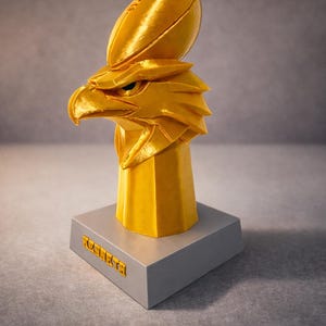 May include: A golden eagle head sculpture with a football on top, mounted on a gray rectangular base. The sculpture has a geometric design and the word "XCONNECTA" is printed on the base.