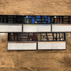 May include: A top-down view of several black plastic trays filled with miniature game pieces. The trays are organized with various colored pieces, including blue, brown, and red. Each tray has a label with text.