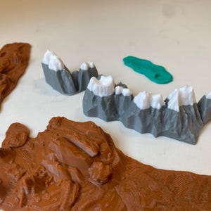 May include: A collection of 3D printed terrain pieces including two brown textured land segments, two gray mountain ranges with white snow-capped peaks, and a small, smooth, oval-shaped green water feature piece for tabletop gaming.