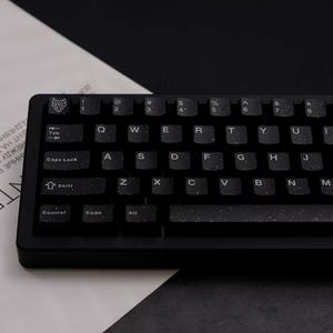 May include: A black mechanical keyboard with white lettering and a wolf logo on the top left corner. The keyboard is on a white paper with text and a black surface.