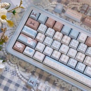 以下が含まれることがあります： パステルブルーのメカニカルキーボード。キーキャップはピンク、ブルー、ホワイトなどのパステルカラーで、リボン、ハート、チェック柄などの装飾が施されています。スペースバーには花の模様があります。可愛らしいデザインです。