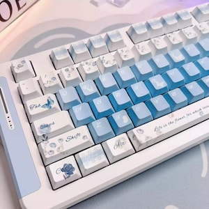Könnte beinhalten: Eine weiße und hellblaue mechanische Tastatur mit blau verlaufenden Tastenkappen. Die Tastenkappen weisen Blumen- und Schmetterlingsdesigns auf, mit dem Text "Life is the flower for which love" auf der Leertaste.
