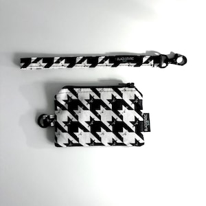 May include: A black and white houndstooth pattern coin purse and wrist strap. The coin purse has a zipper closure and a small black ring. The wrist strap has a black clasp. Both items have a repeating cat face pattern.