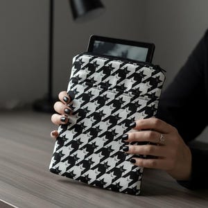 May include: Black and white houndstooth patterned tablet sleeve with a zipper closure. The sleeve is holding a black tablet. The sleeve is being held by a person with black nail polish.