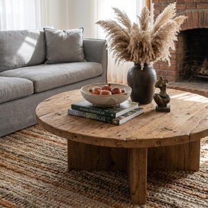 May include: A rustic, round wooden coffee table with a natural finish. The table holds a ceramic bowl of pomegranates, two books, a dark vase with dried pampas grass, and a small bronze sculpture. A gray sofa and patterned rug complete the living room scene.