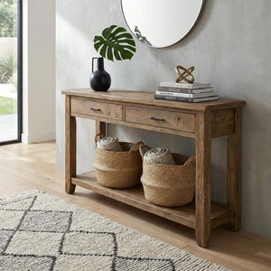 May include: A wooden console table with two drawers and a lower shelf, holding two woven baskets. The table is topped with a black vase, a green leaf, books, and a gold sculpture. A round mirror hangs on the wall behind the table.