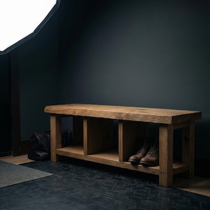 Puede incluir: Banco de madera r&uacute;stico con acabado natural. El banco tiene un estante inferior y dos compartimentos abiertos. Un par de botas de cuero marr&oacute;n est&aacute;n en el estante. El banco est&aacute; frente a una pared oscura.
