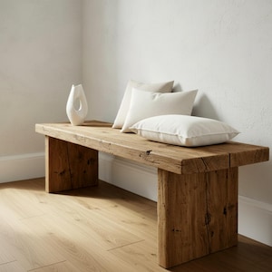 Puede incluir: Banco de madera r&uacute;stico con acabado natural. El banco tiene una parte superior rectangular gruesa y patas robustas. Tres cojines blancos descansan sobre el banco y un jarr&oacute;n blanco est&aacute; a la izquierda. El banco est&aacute; contra una pared blanca.