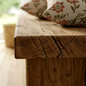Puede incluir: Primer plano de un banco de madera r&uacute;stico con vetas y grietas visibles. Dos cojines decorativos con estampados florales descansan sobre el banco. El banco tiene un aspecto natural y envejecido, lo que sugiere que est&aacute; hecho de madera recuperada.