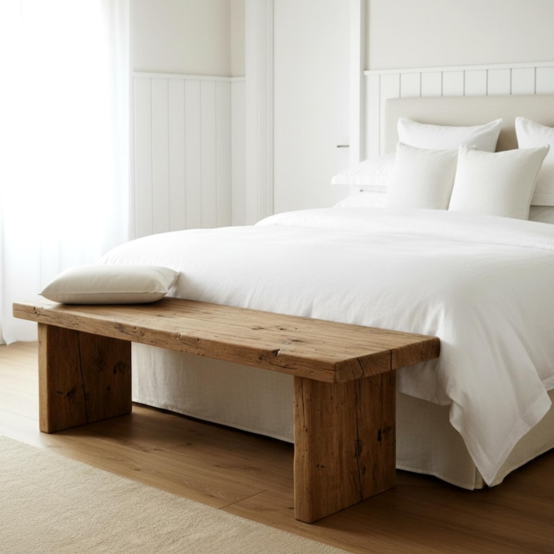 Puede incluir: Banco de madera r&uacute;stica con acabado natural. El banco es rectangular, con patas gruesas y una parte superior s&oacute;lida. Una almohada blanca descansa sobre el banco. El banco est&aacute; en un dormitorio con una cama y ropa de cama blancas.
